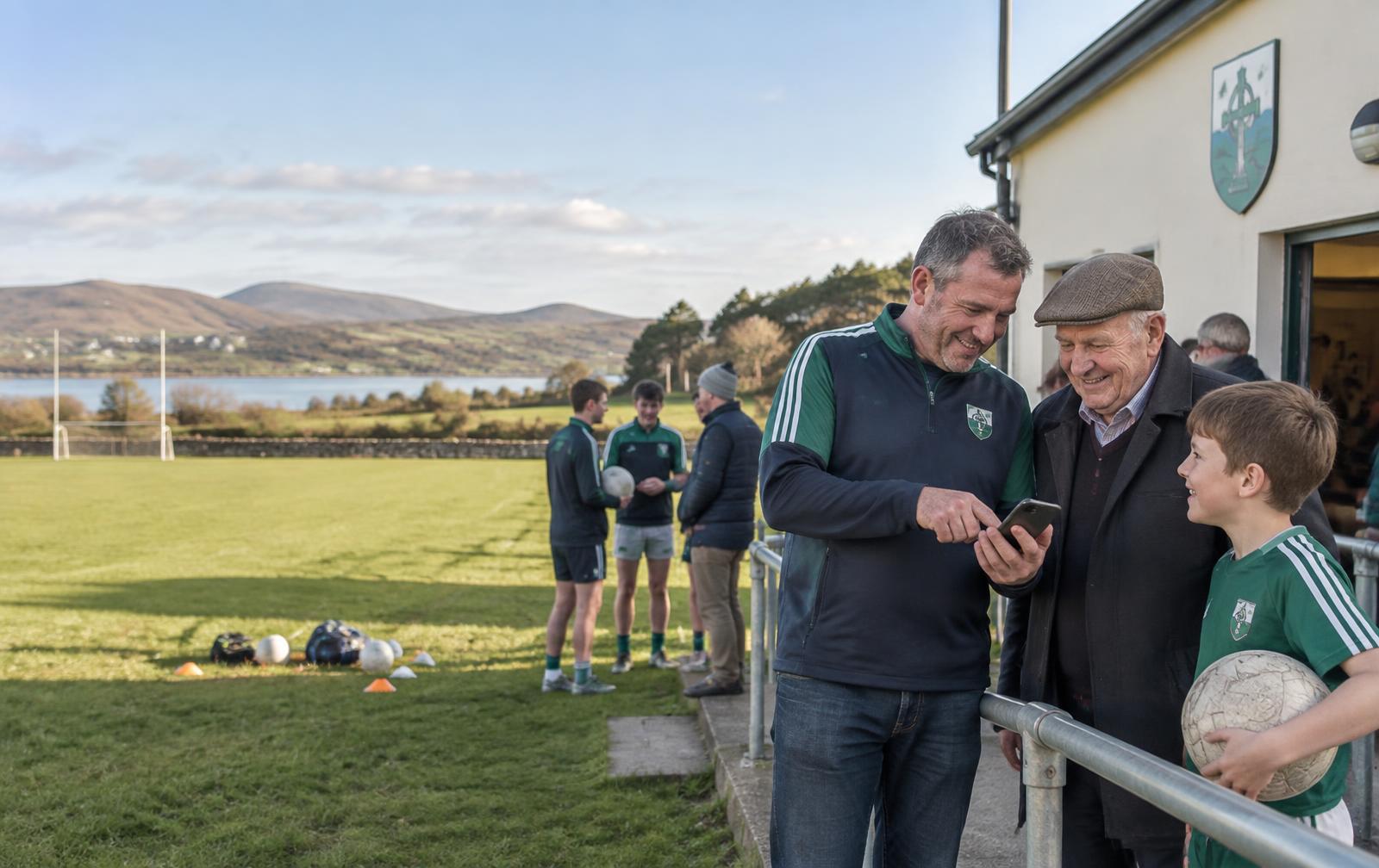 Club volunteer helping an older member on a phone beside a GAA pitch